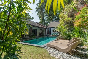 A swimming pool surrounded by lush greenery, a lounge chair, and a small patio area.