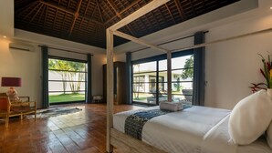 A spacious bedroom with a large bed, wooden flooring, and a view of a lush garden through large windows.
