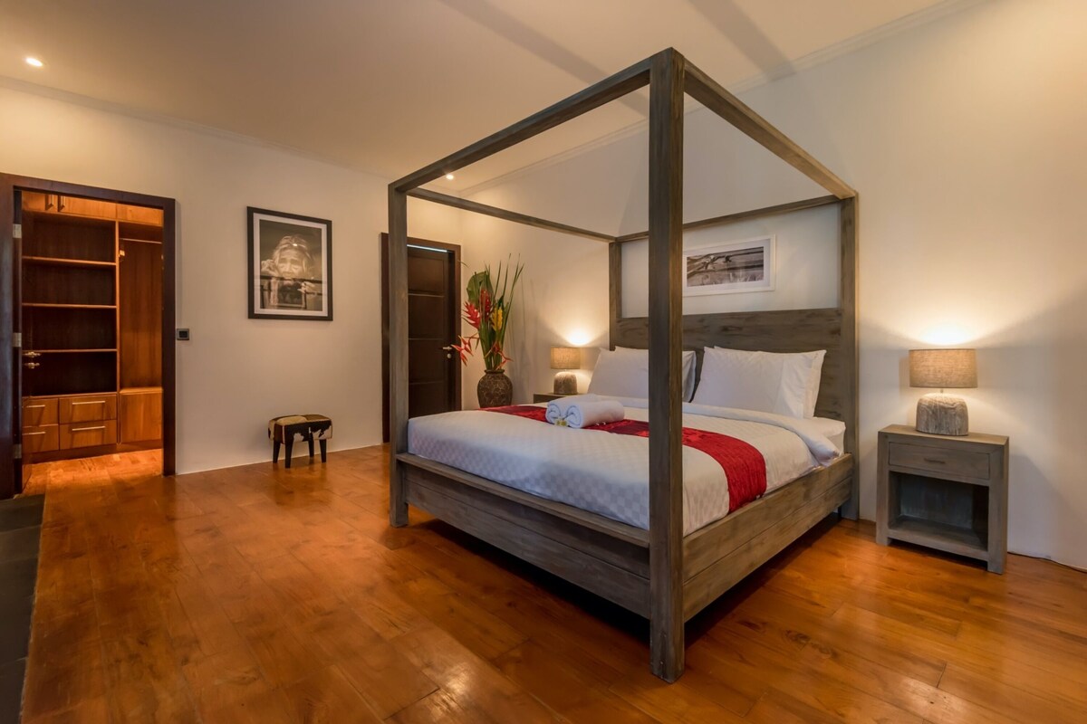 A four-poster bed with a wooden frame, a bedside table with a lamp, and a small stool.