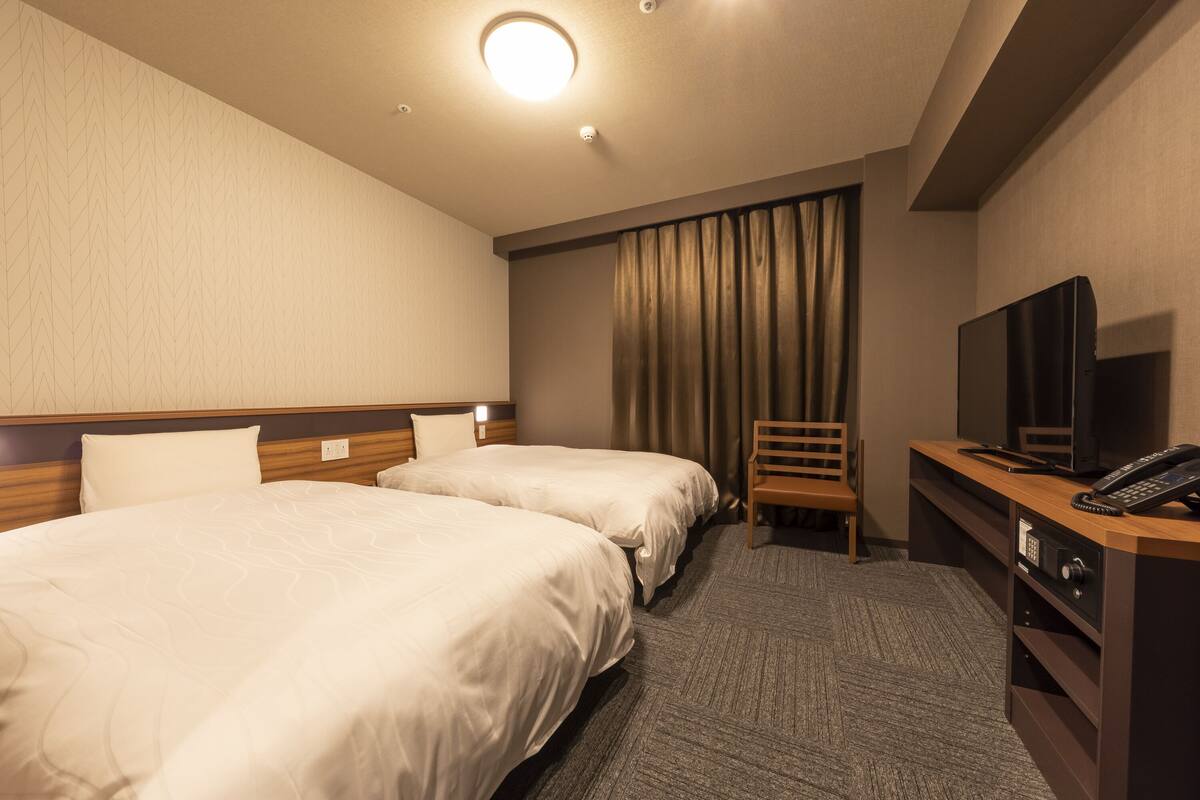 A hotel room with two beds, a desk with a television and phone, a chair, and a window with curtains.