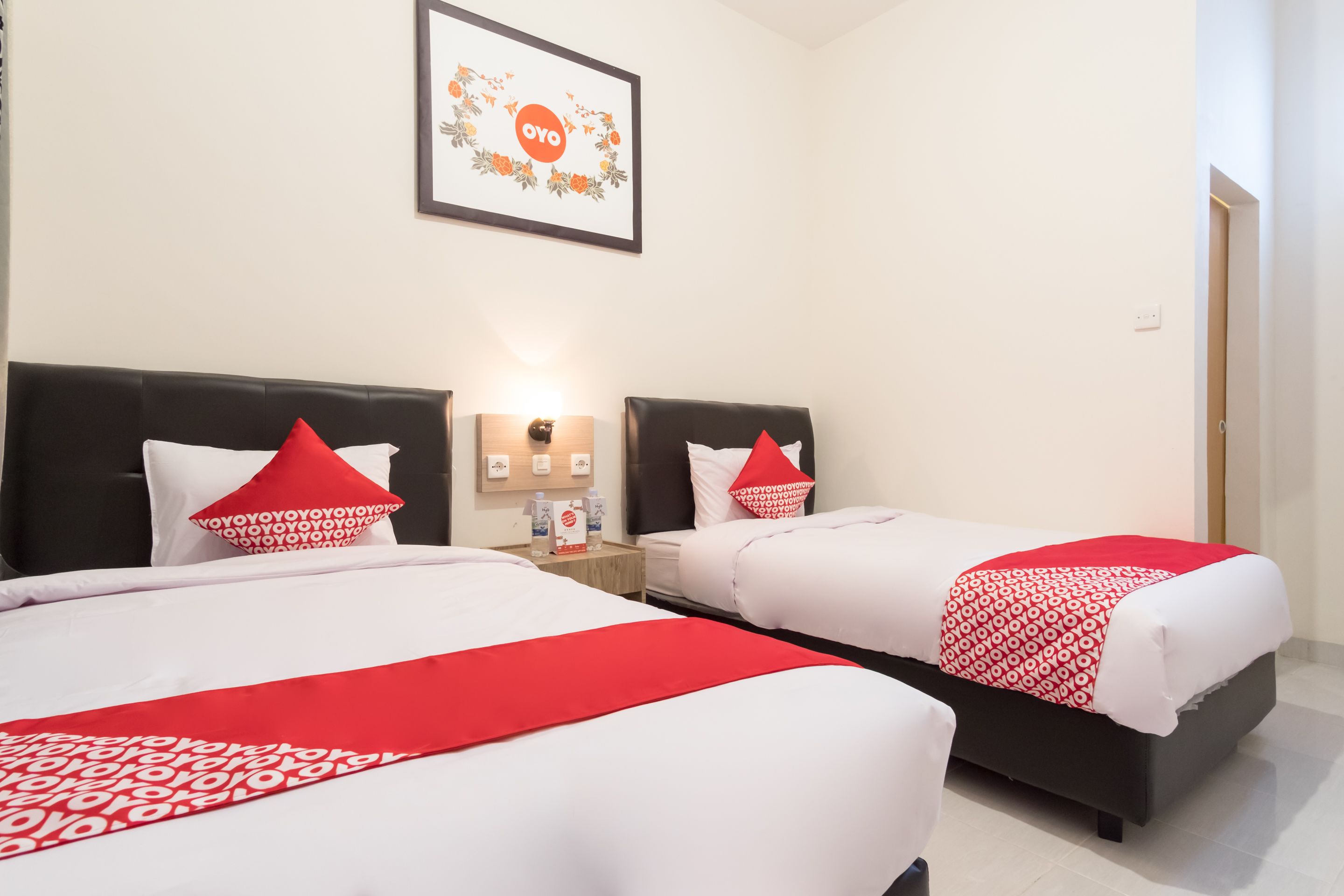 Two beds with red and white bedding, a framed picture on the wall, and a nightstand with a lamp and water bottles.