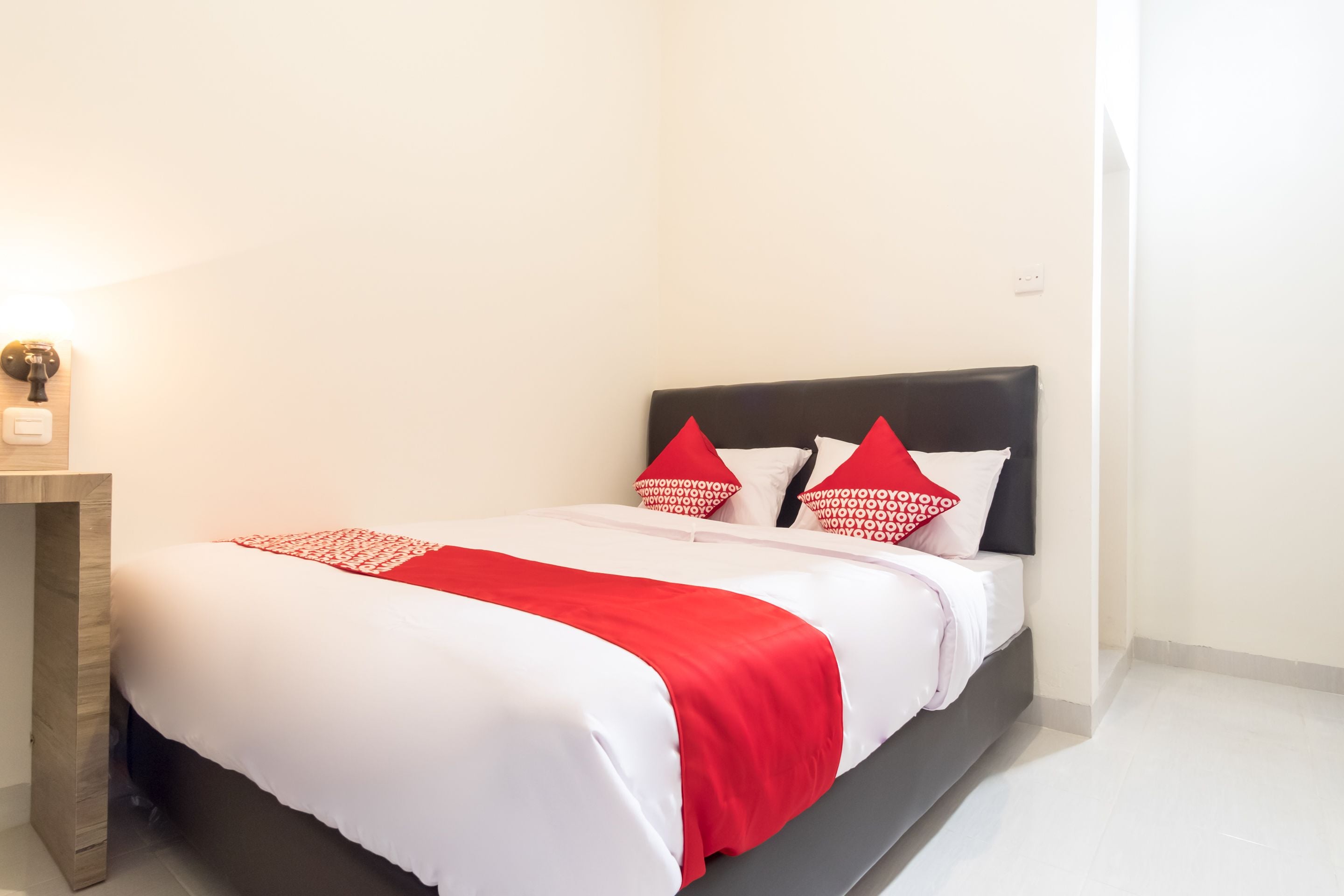 A neatly made bed with white and red bedding, a dark headboard, and a bedside table with a lamp.