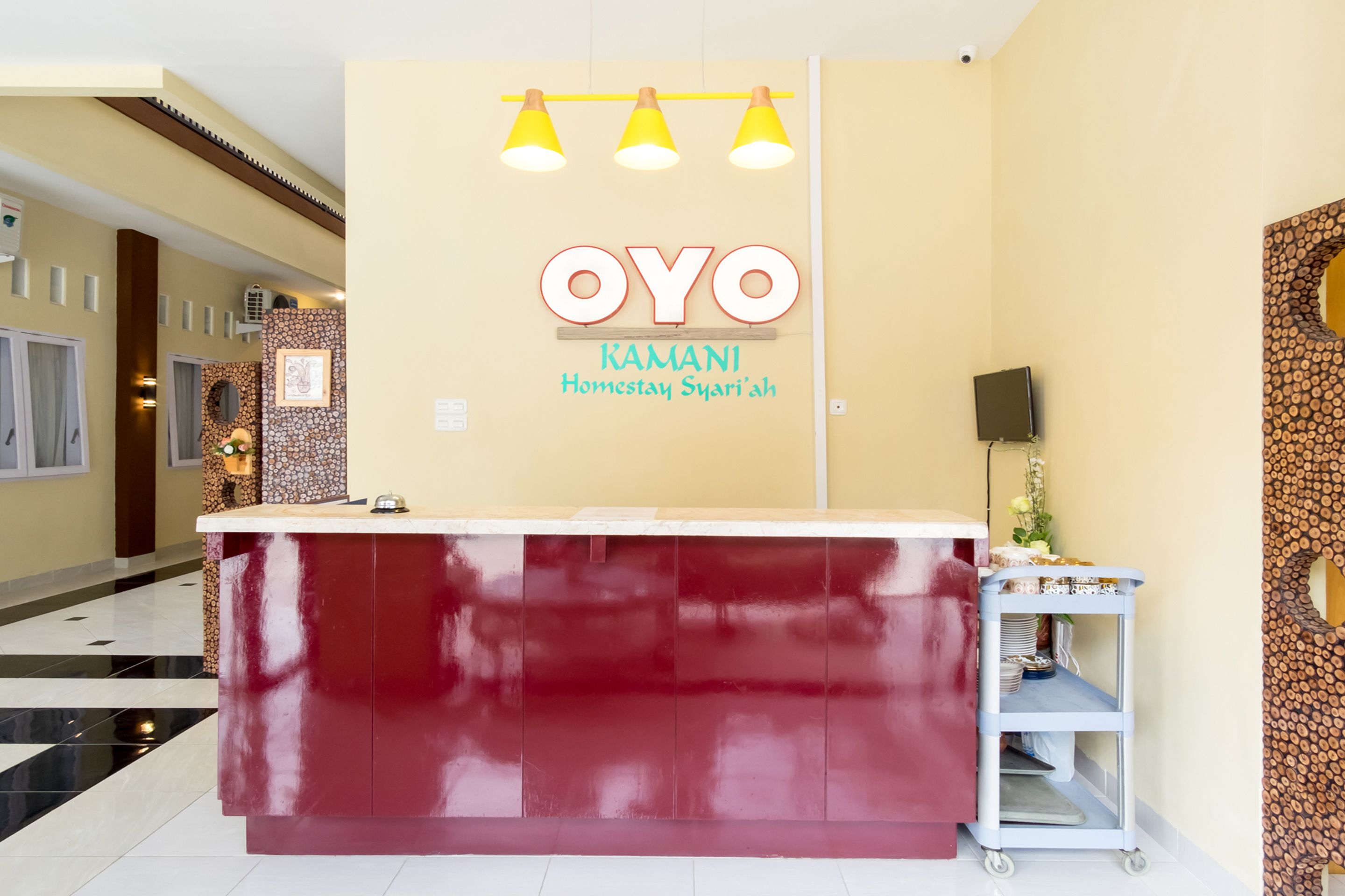 Ein Empfangsbereich mit einem dunkelroten Tresen, einem Schild mit der Aufschrift 'OYO KAMANI Homestay Syari'ah' und einem an der Wand befestigten Fernseher.