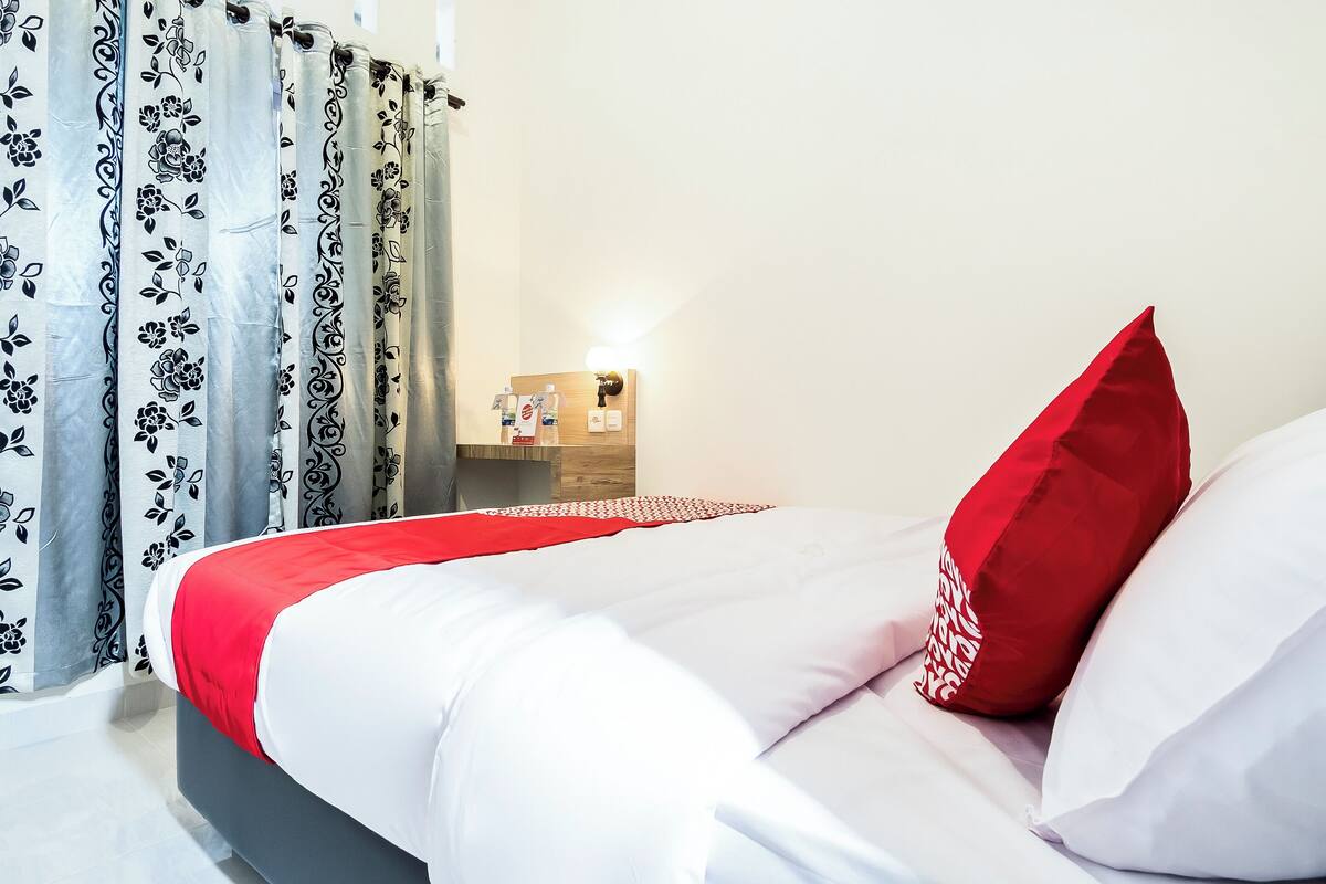 A neatly made bed with white and red bedding, a red pillow, and a grey and black floral curtain.