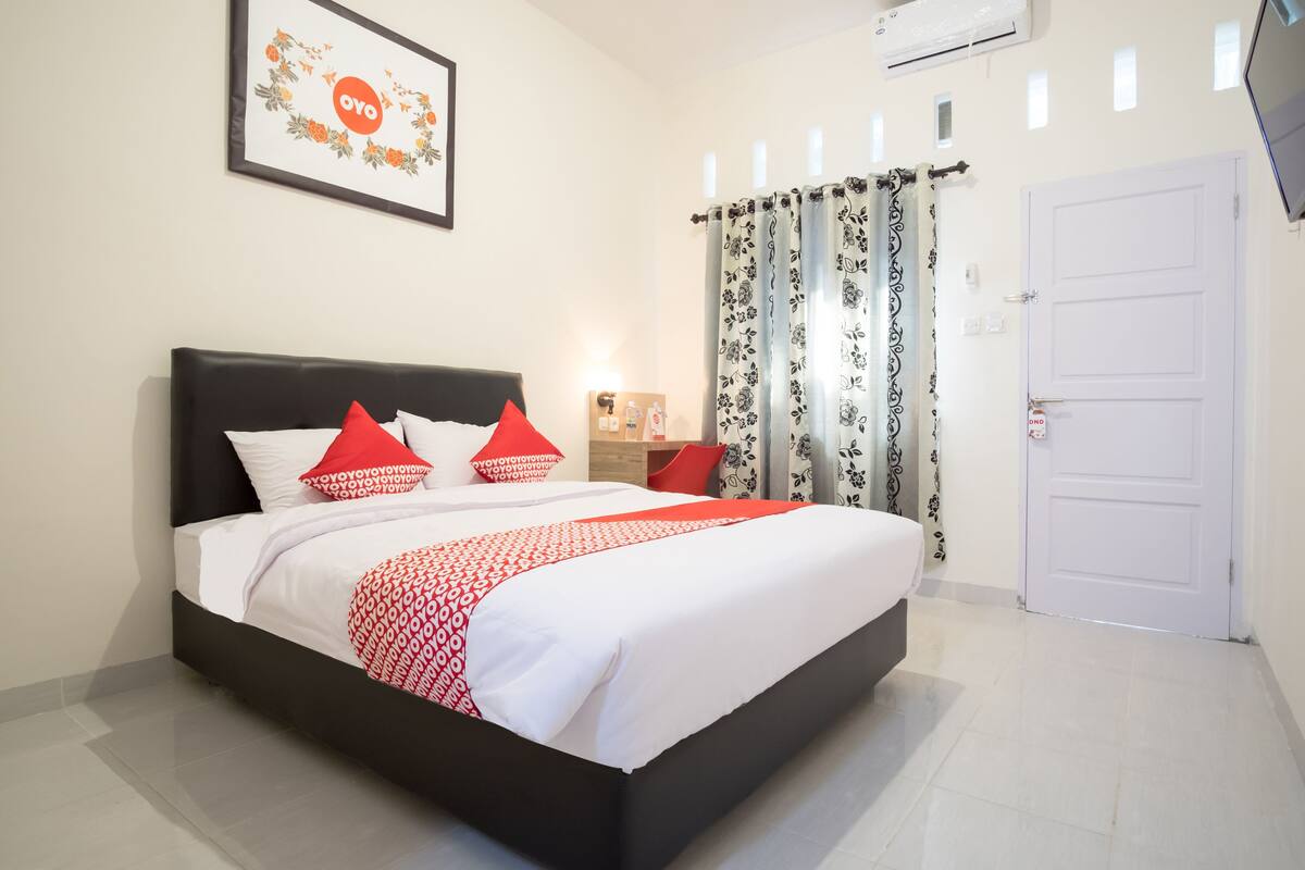 A neatly made bed with a patterned bedspread and pillows, a bedside table with a lamp, and a door with a security lock.