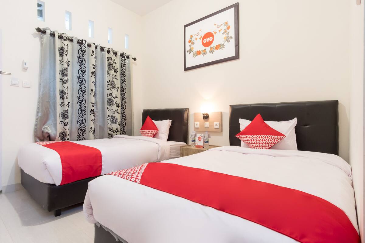 A hotel room with two beds, each with a red and white bedspread, black headboards, and matching red pillows. There is a framed picture on the wall, a nightstand with a lamp, and a window with patterned curtains.
