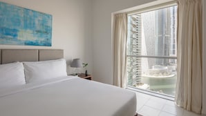 A hotel room with a large bed, a nightstand with a lamp, a window with curtains, and a view of high-rise buildings. - Fantastay Marina Tower (Dubai)