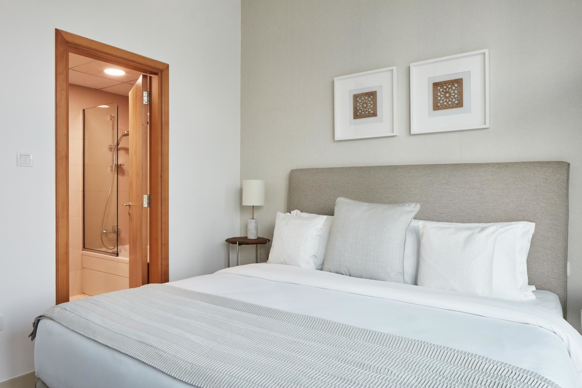 A hotel room with a bed, a wooden door, a nightstand with a lamp, and two framed pictures on the wall.