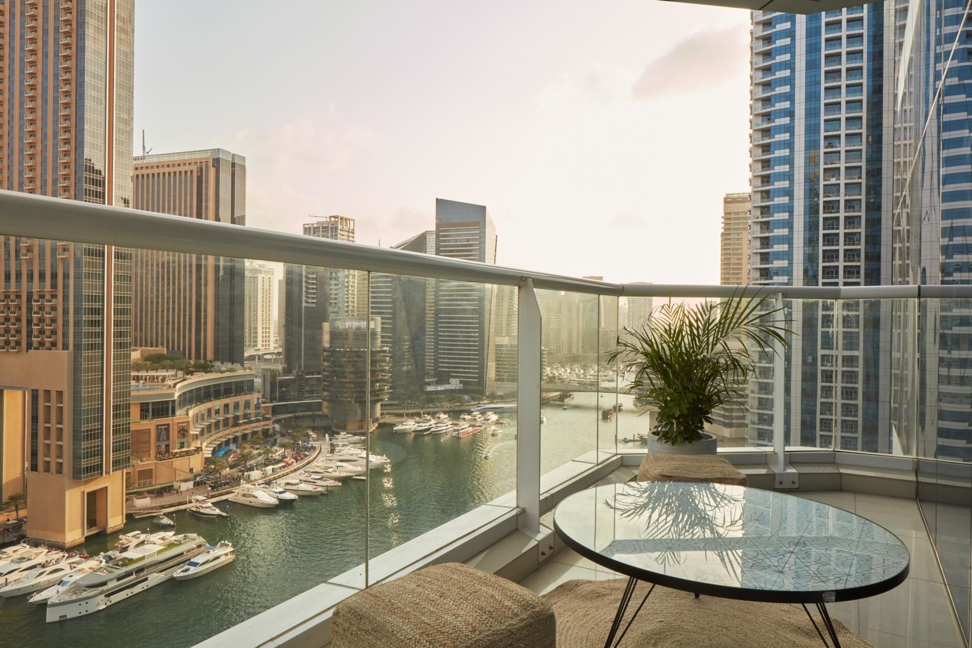 A balcony with a glass table and a potted plant, overlooking a marina with boats and modern buildings.
