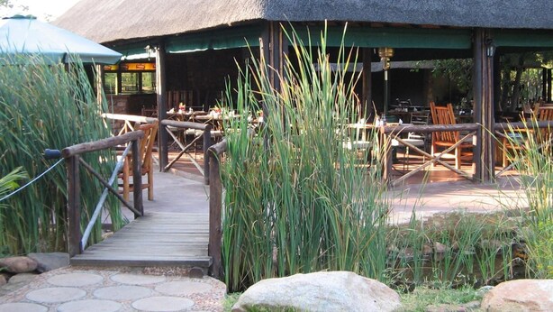 A restaurant with a thatched roof, outdoor seating, and a wooden pathway leading to the entrance. - Phokoje Bush Lodge (Selebi-Phikwe)