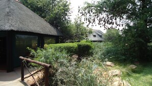 A thatched-roof building with a wooden deck, surrounded by dense greenery and a rock garden. - Phokoje Bush Lodge (Selebi-Phikwe)