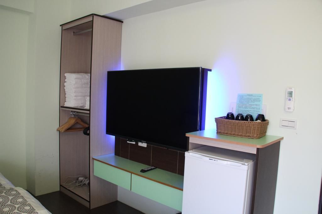 A compact hotel room with a flat-screen TV mounted on the wall, a small refrigerator, a wooden wardrobe with hanging clothes, and a basket of toiletries on a small table.
