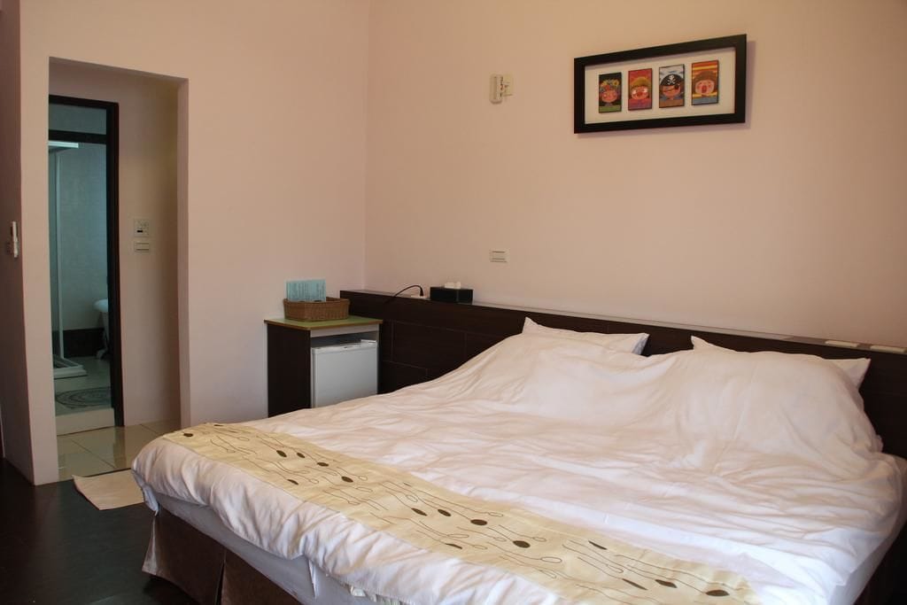 A hotel room with a bed, a small bedside table, a framed picture on the wall, and a door leading to another room.