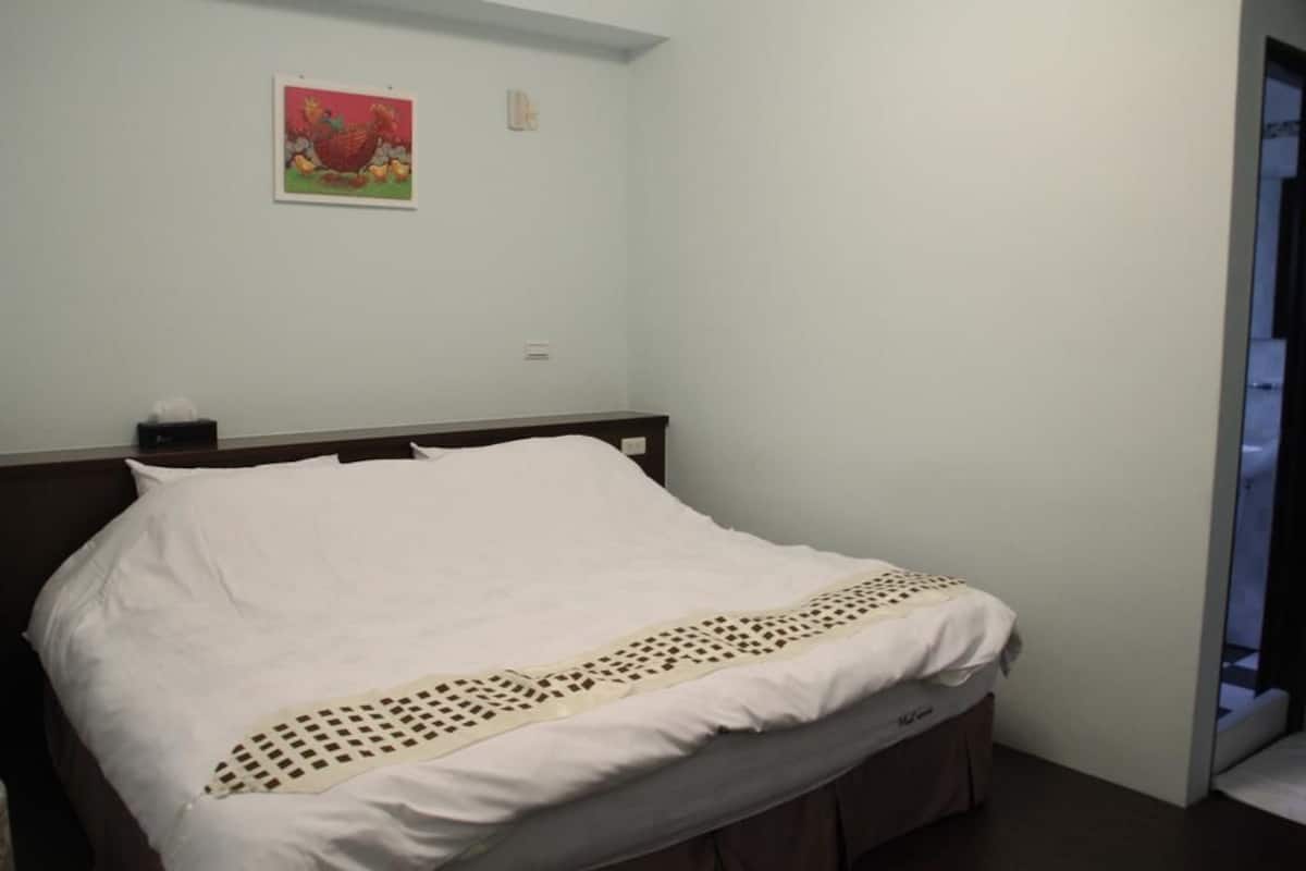A single bed with a white bedspread and a decorative border, a wooden headboard, and a wall-mounted light switch.