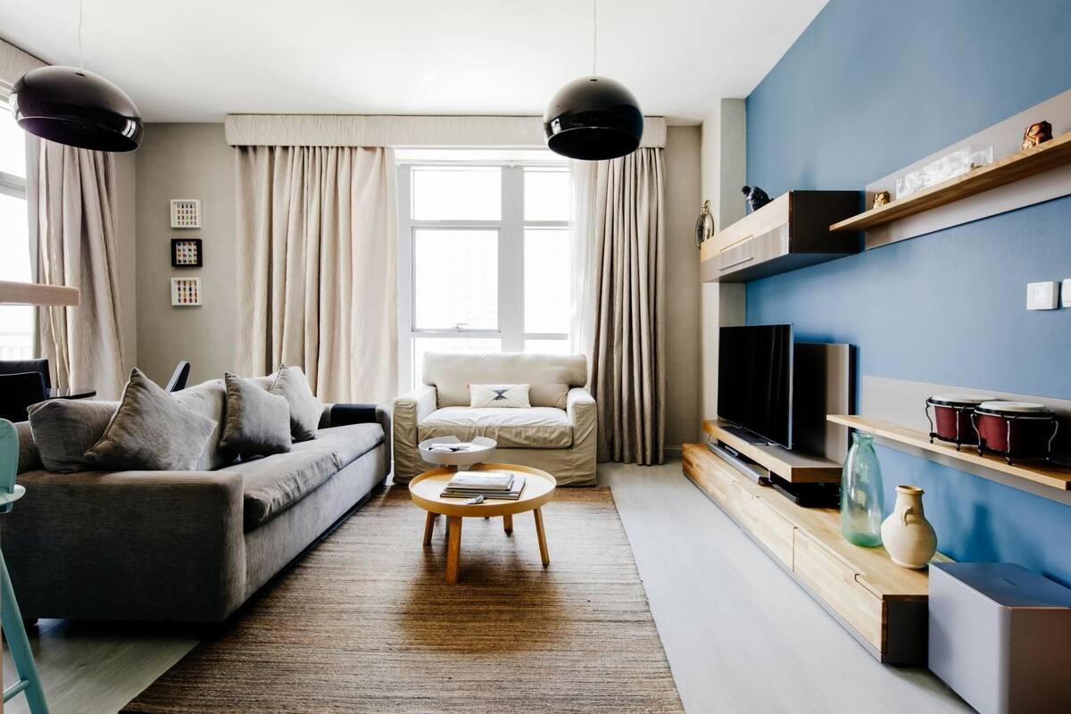A modern living room with a sofa, armchair, coffee table, and a TV. There are curtains, a window, and a shelf with decorative items.