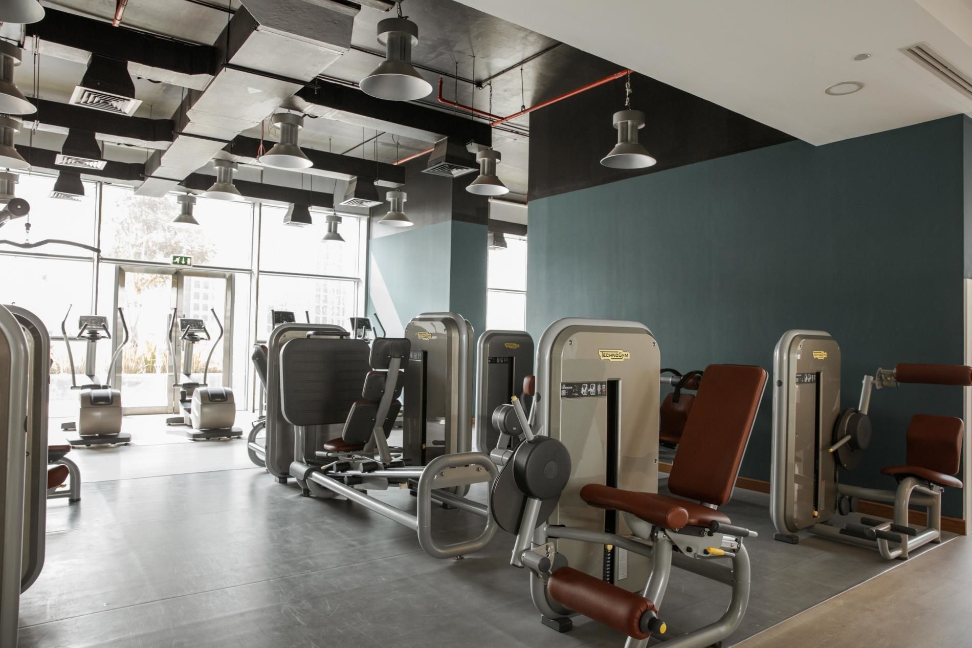 A gym with various exercise machines, large windows, and a modern ceiling design.