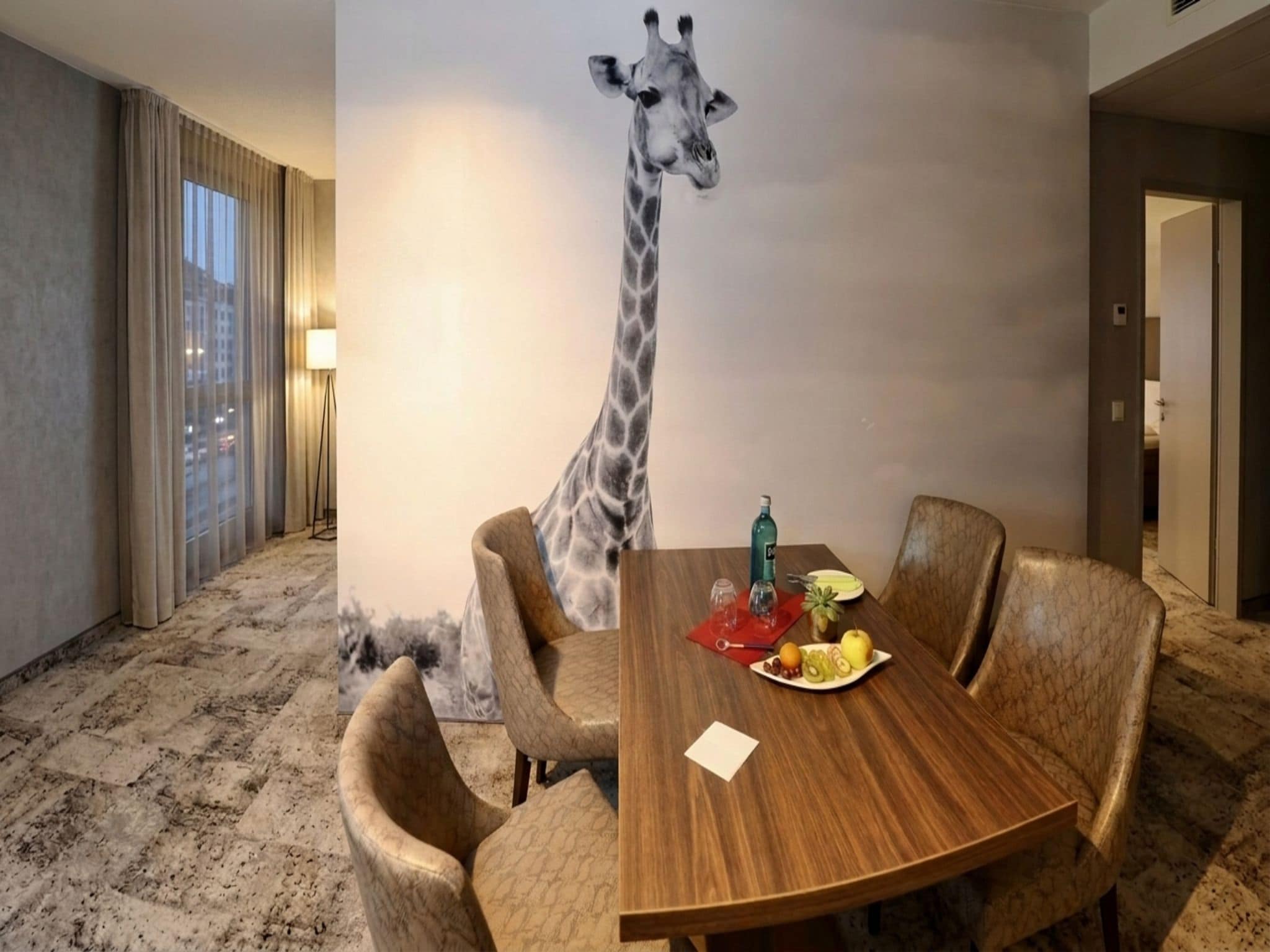 Habitación de hotel con mesa de comedor, sillas, un mural de jirafa y vista a la ciudad por la ventana.