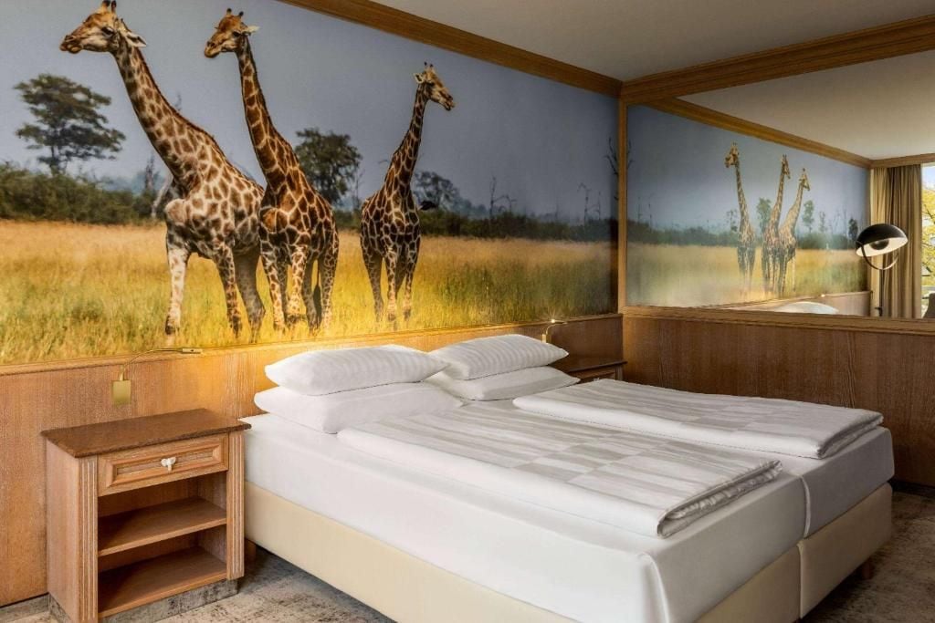 Habitación de hotel con un gran mural de jirafas, una cama con sábanas blancas, una mesita de noche y una lámpara.
