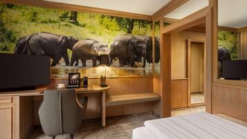 Una habitación de hotel con escritorio, cama, televisor y un mural de elefantes.