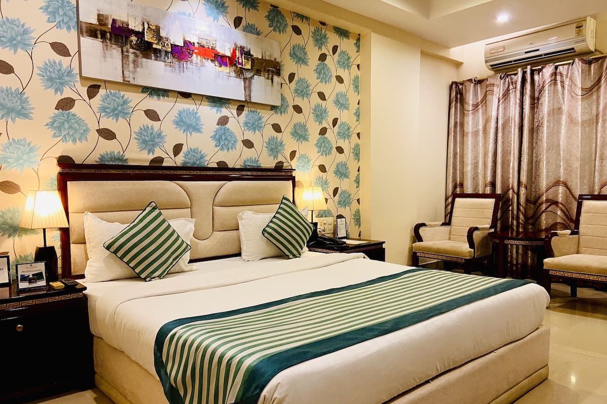 A hotel room with a large bed, two bedside tables, a nightstand, a chair, a desk, a wall-mounted air conditioner, and a decorative wall piece.