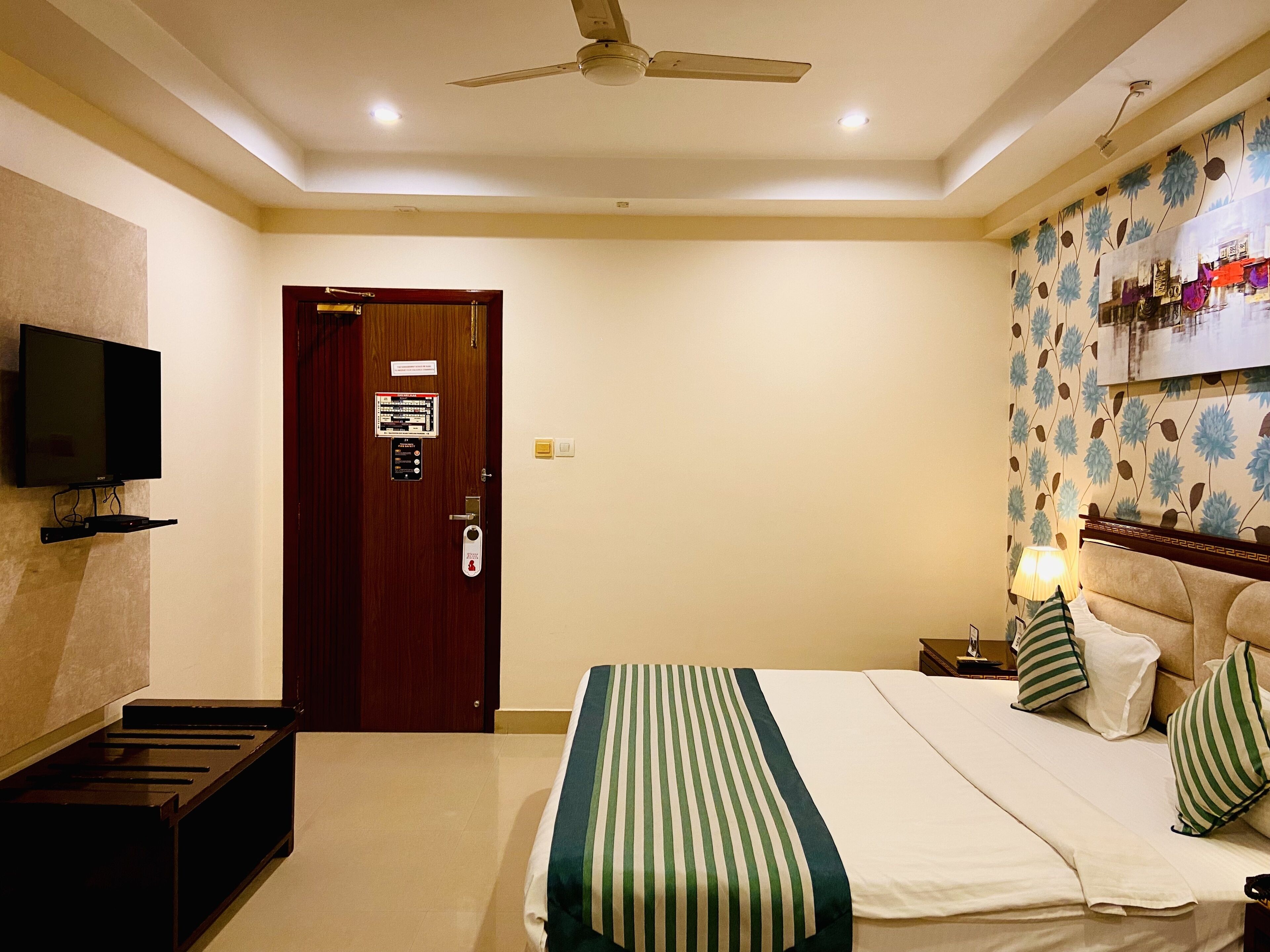 A hotel room with a bed, a television mounted on the wall, a ceiling fan, and a door with a sign.