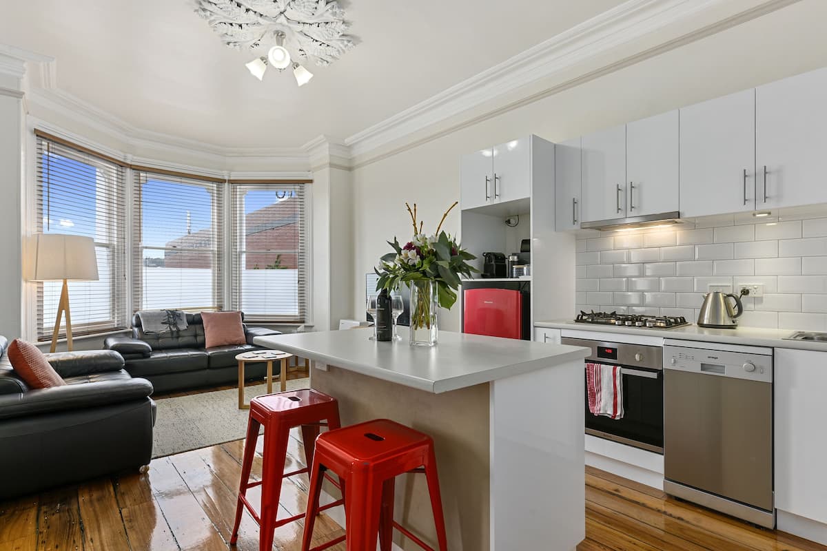 A modern kitchen with white cabinets, a central island, red stools, and a view through large windows.