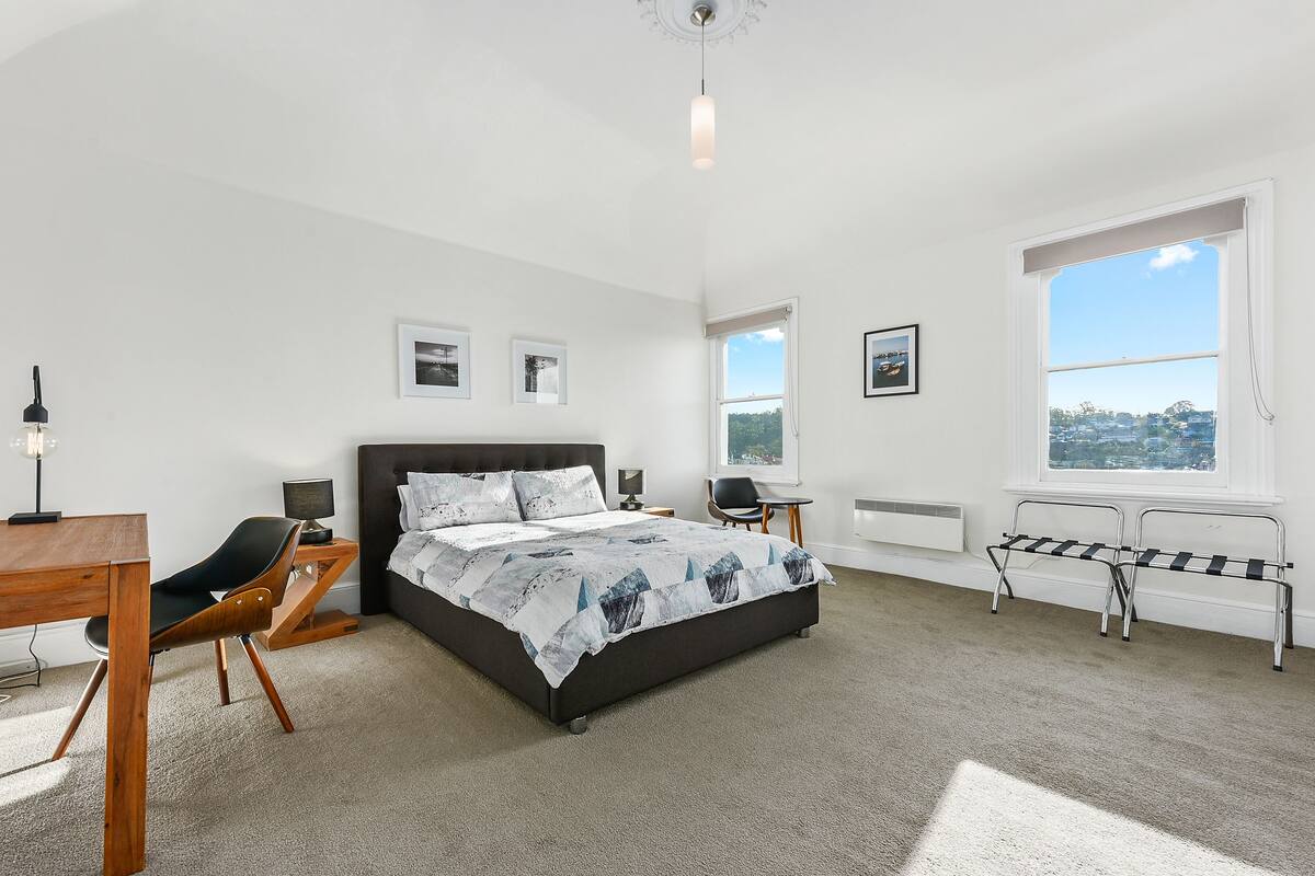 A bedroom with a bed, a desk, a chair, a bench, and a window with a view.