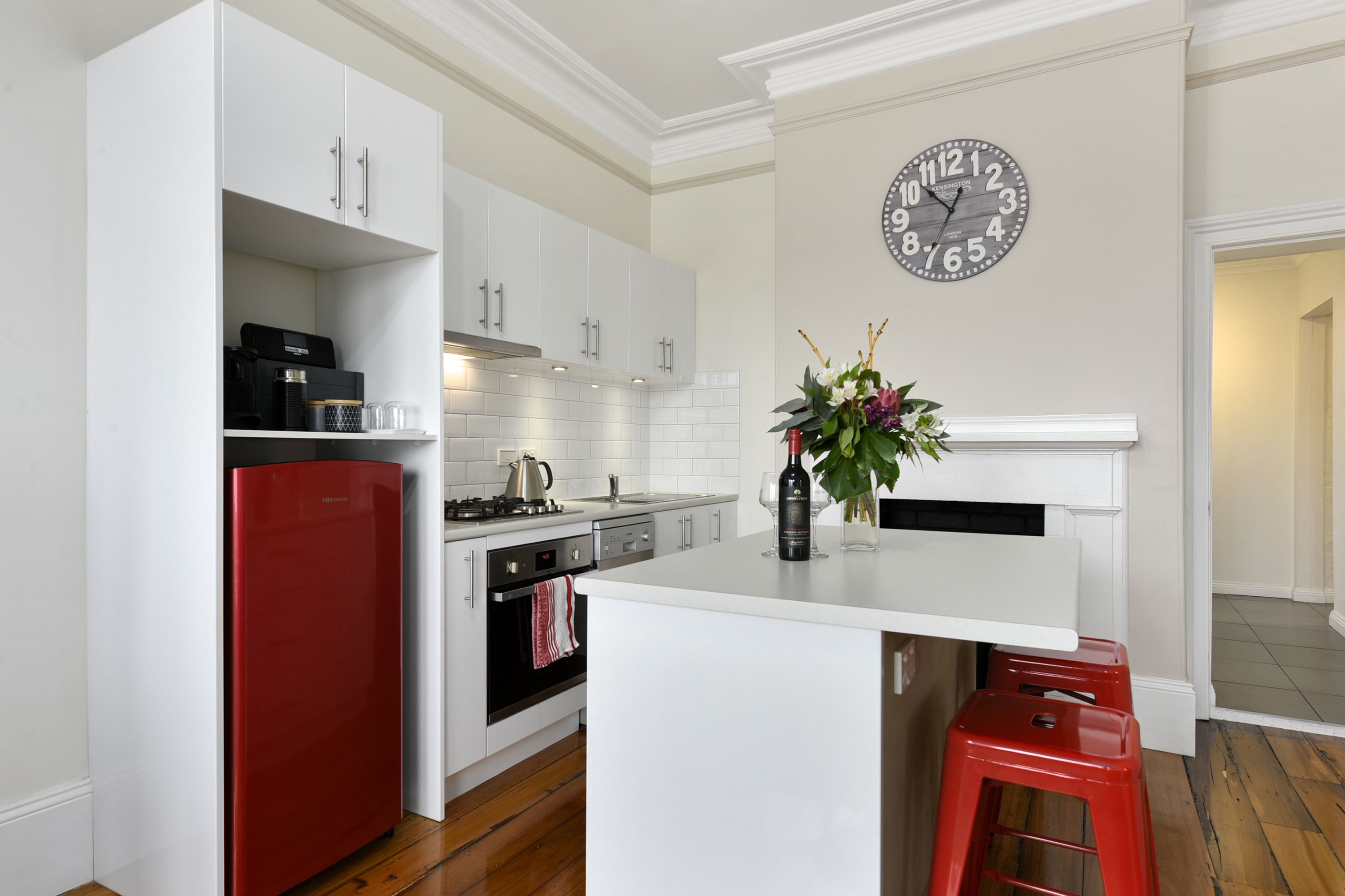 A modern kitchen with white cabinetry, a red refrigerator, a built-in oven, and a clock on the wall.