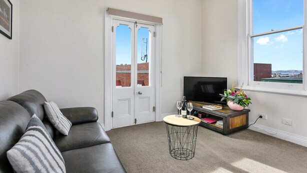 A living room with a leather sofa, a TV, a small round table with drinks, and a window with a view of buildings. - Trinity Hill Apartments (Hobart)
