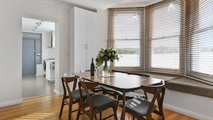 A dining area with a wooden table and chairs, a vase with flowers, and a view through large windows with blinds. - Trinity Hill Apartments (Hobart)