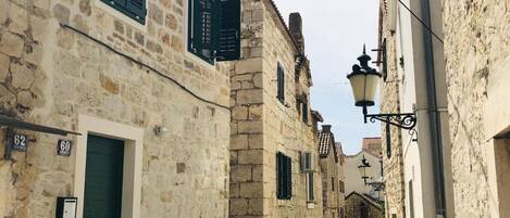 A narrow street lined with stone buildings, featuring traditional architecture with shutters and a clear sky above.