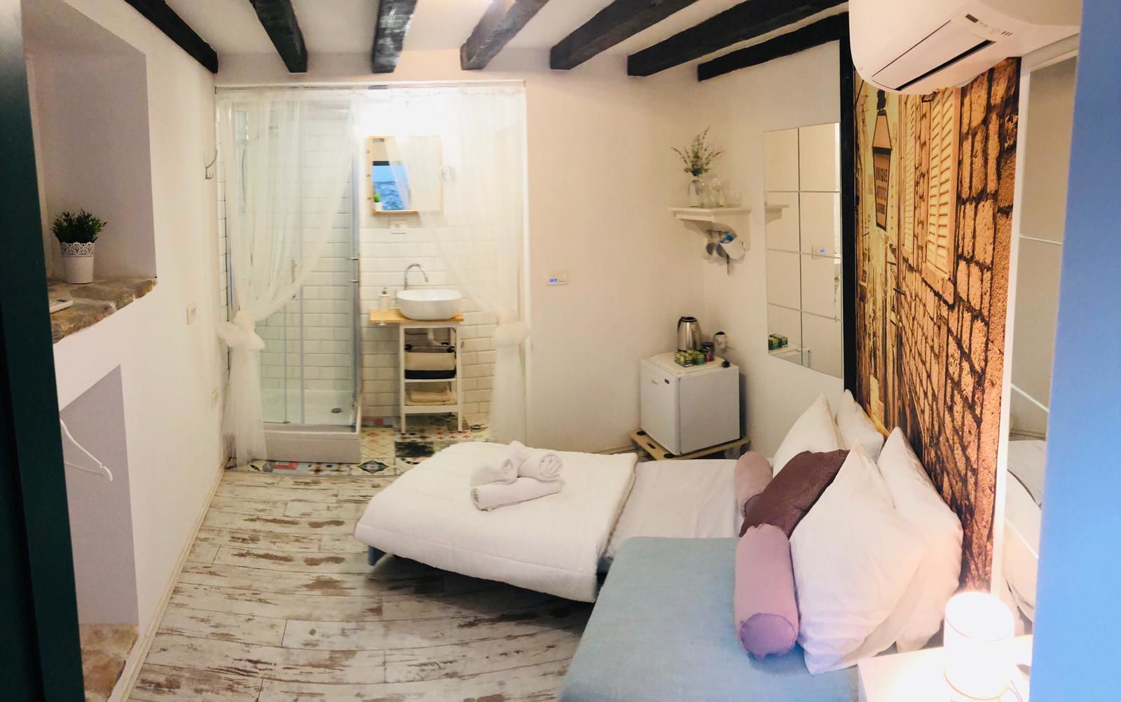 A small, compact bedroom with a bed, a bedside table, a mirror, and a small shelf with a plant.