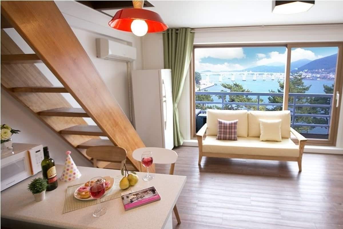 A modern room with a wooden staircase, a sofa with cushions, a dining table with food and drinks, and a large window with a view of the sea and mountains.