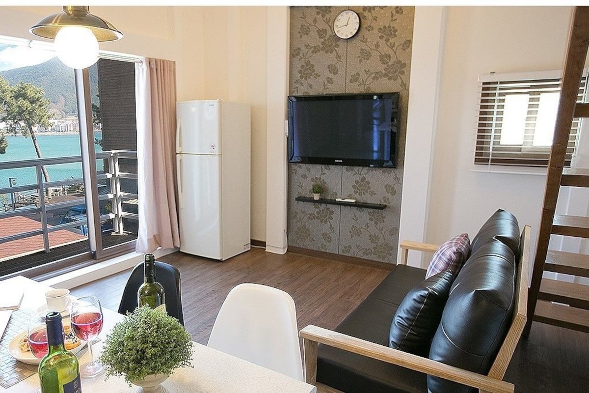 A room with a balcony, a dining area with a table set for two, a sofa, a TV mounted on the wall, and a refrigerator.