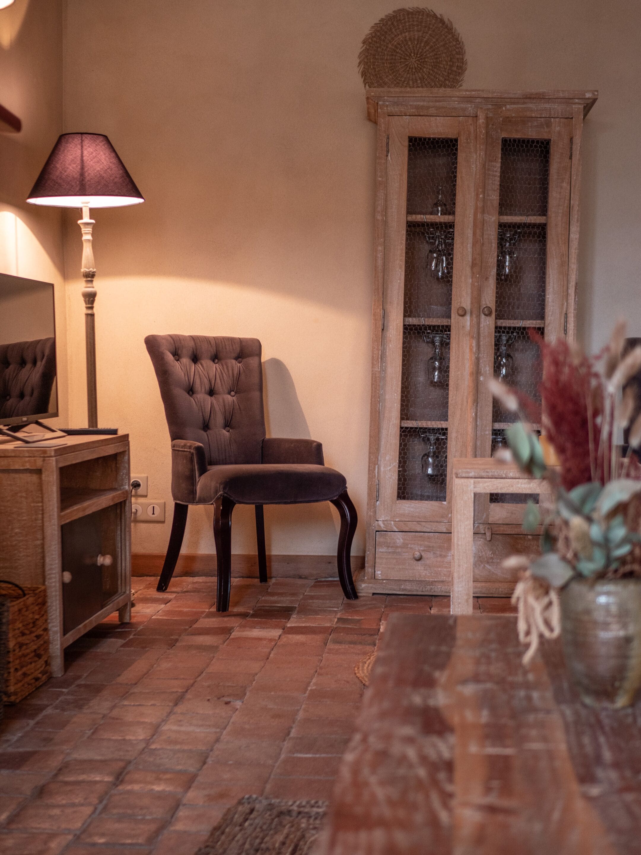 A cozy room with a tufted armchair, a wooden cabinet with glass doors, a lamp, and a vase with dried flowers.