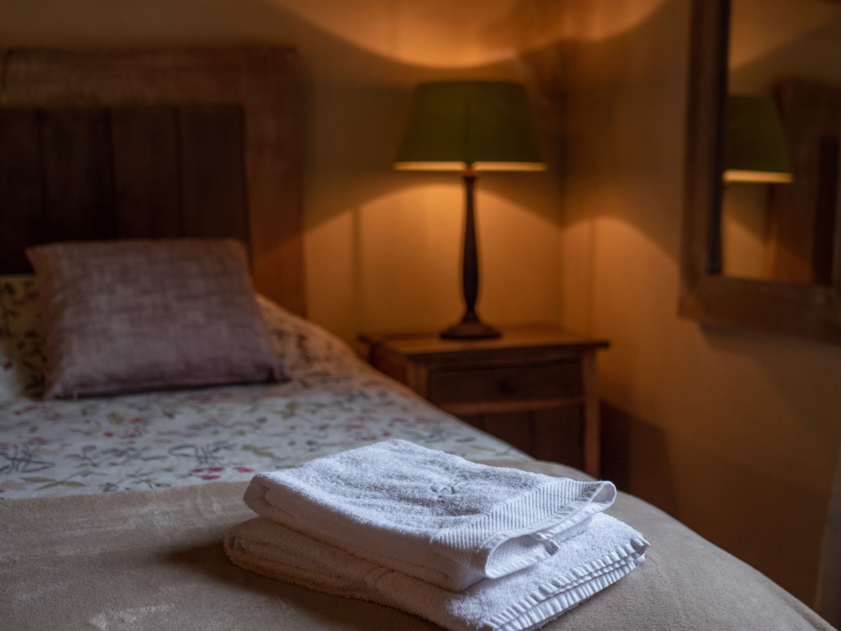 Una cama bien hecha con una toalla blanca doblada, una lámpara verde en una mesita de noche y un cojín estampado con motivos florales.