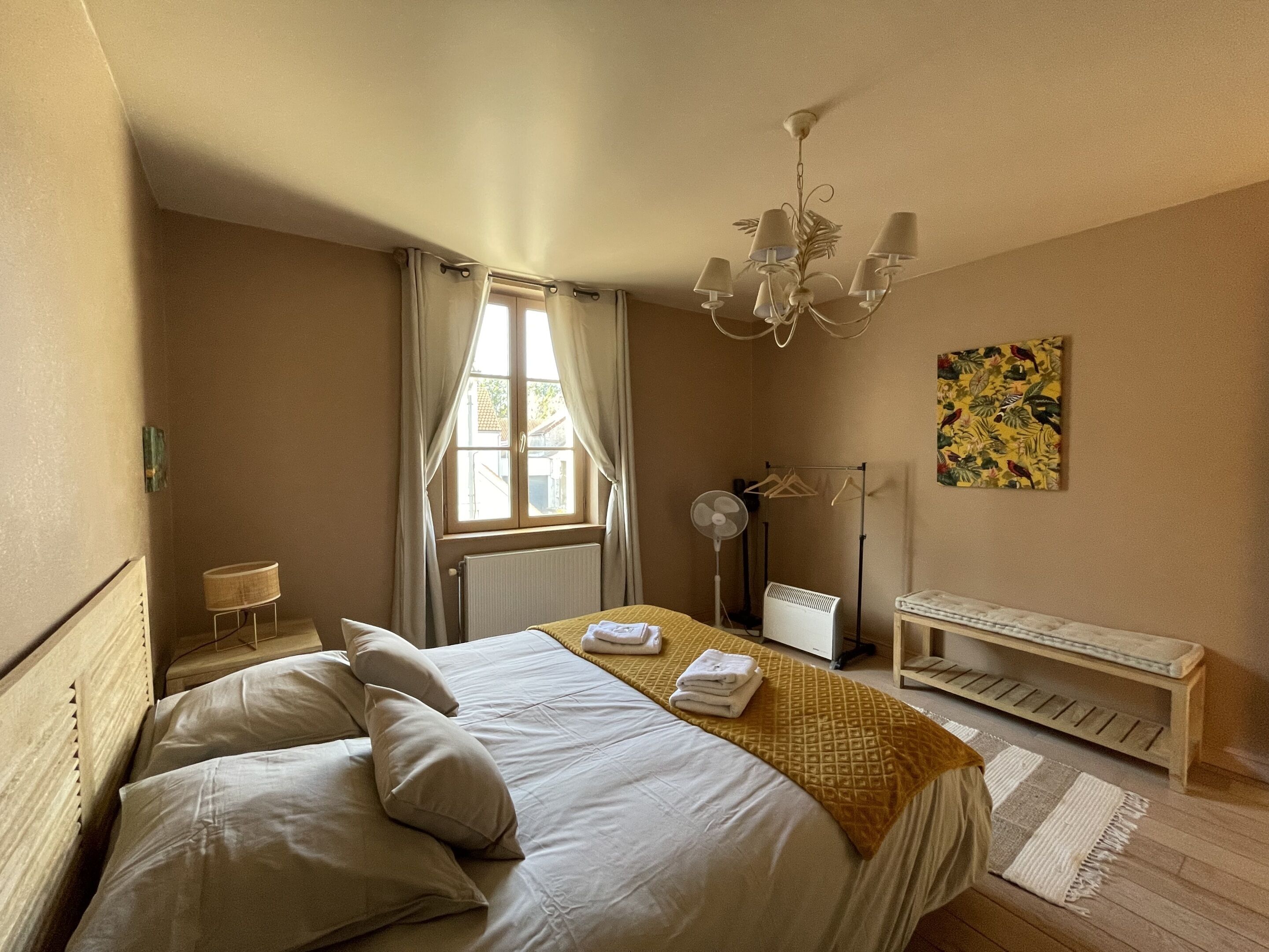 Hotelkamer met een bed, nachtkastje, lamp, radiator, ventilator, een bankje en een raam met gordijnen.