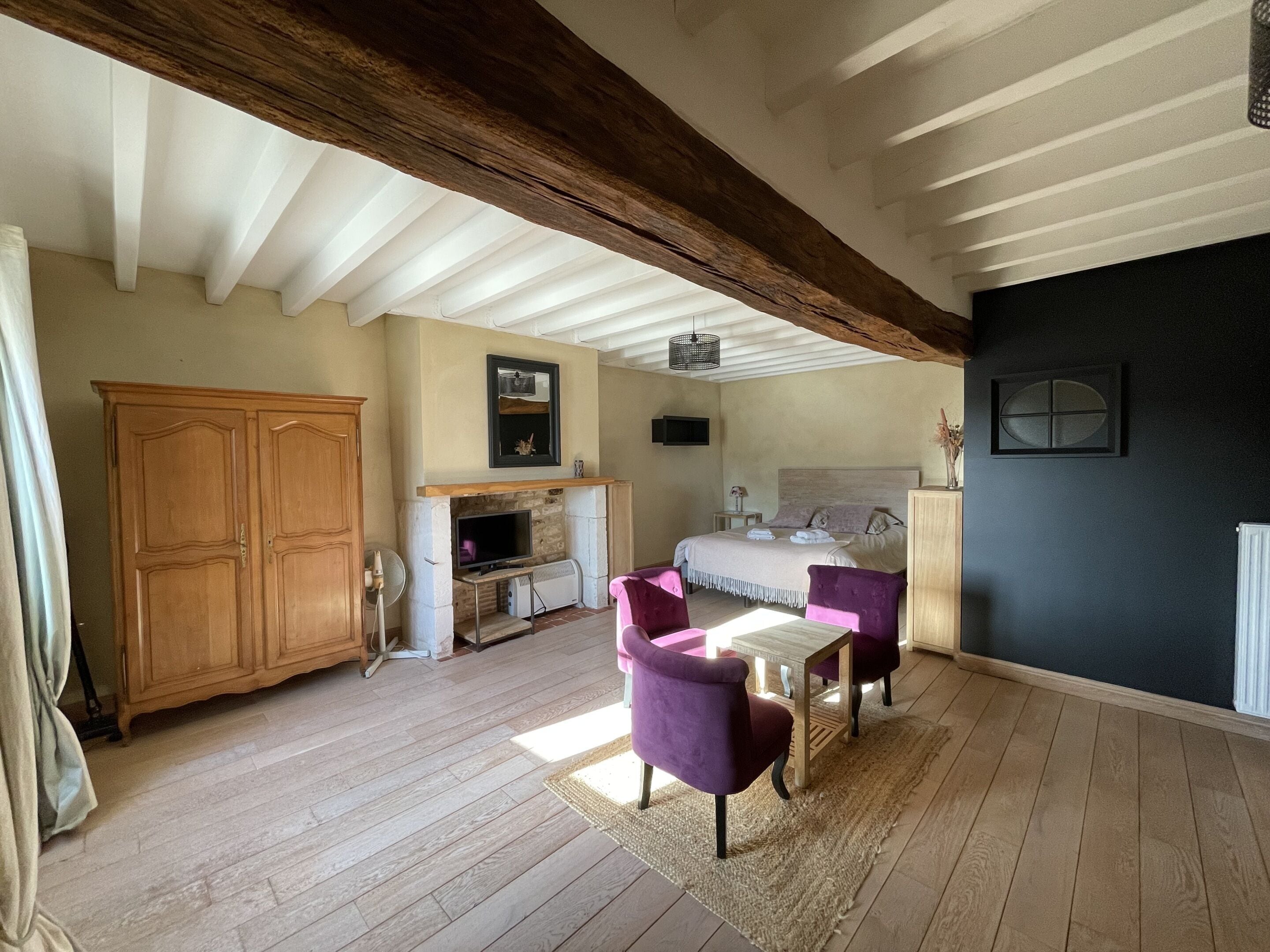 A spacious room with a large wooden wardrobe, a fireplace, a bed with a headboard, and a dining area with two purple chairs.