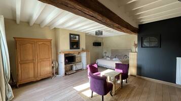 A spacious room with a large wooden wardrobe, a fireplace, a bed with a headboard, and a dining area with two purple chairs.