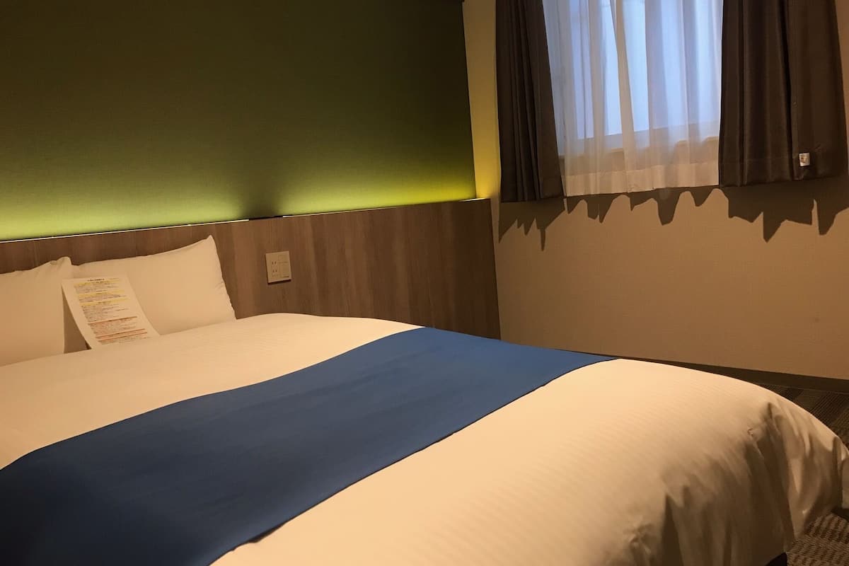 Una habitación de hotel con una cama, un cabecero verde, una ventana con cortinas y un interruptor de luz.