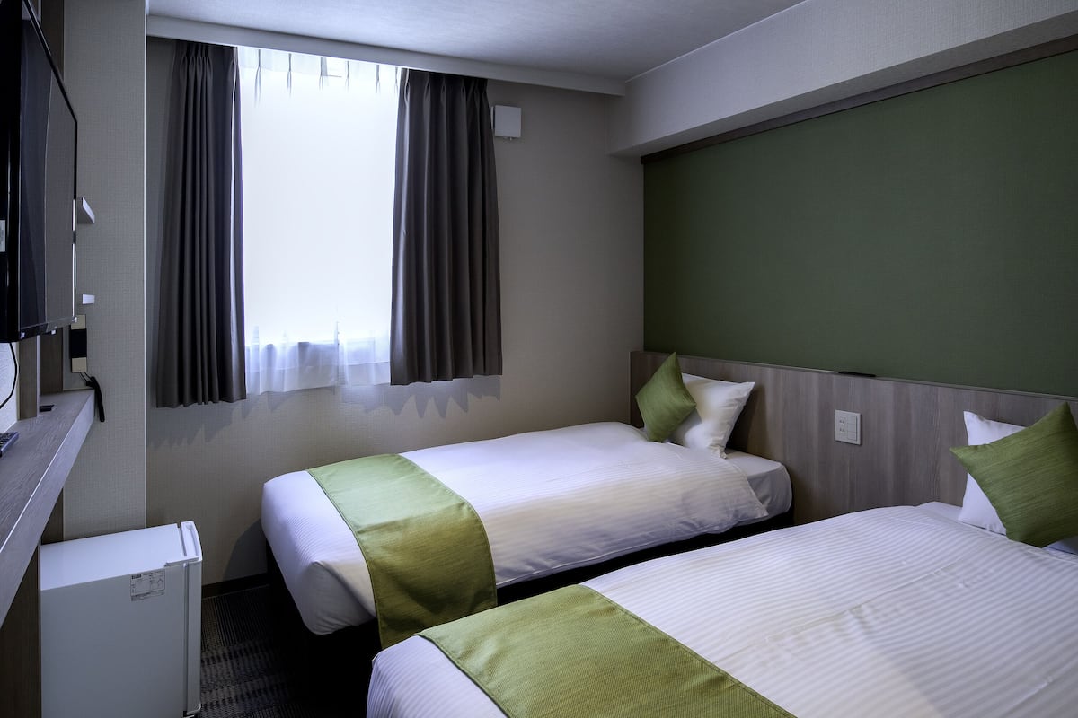 Habitación de hotel con dos camas, un refrigerador pequeño, un escritorio con teléfono y una ventana con cortinas.