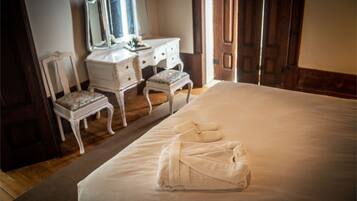 A neatly made bed with a folded towel, a white dresser with a mirror, and a chair with a floral cushion in a hotel room.