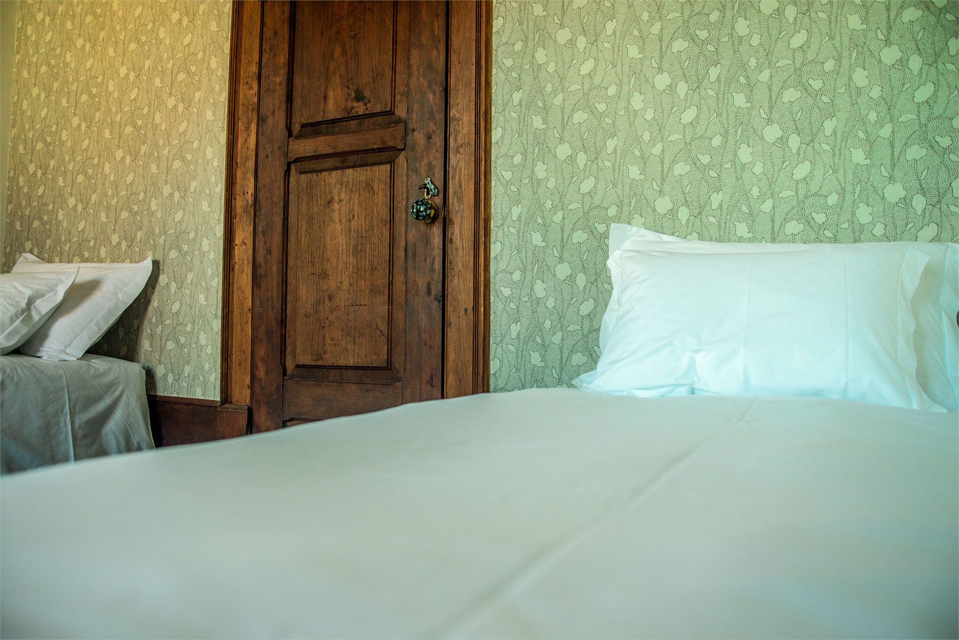 Ein Zimmer mit Holztür, einem Bett mit weißer Bettwäsche und Wandtapete mit Muster.