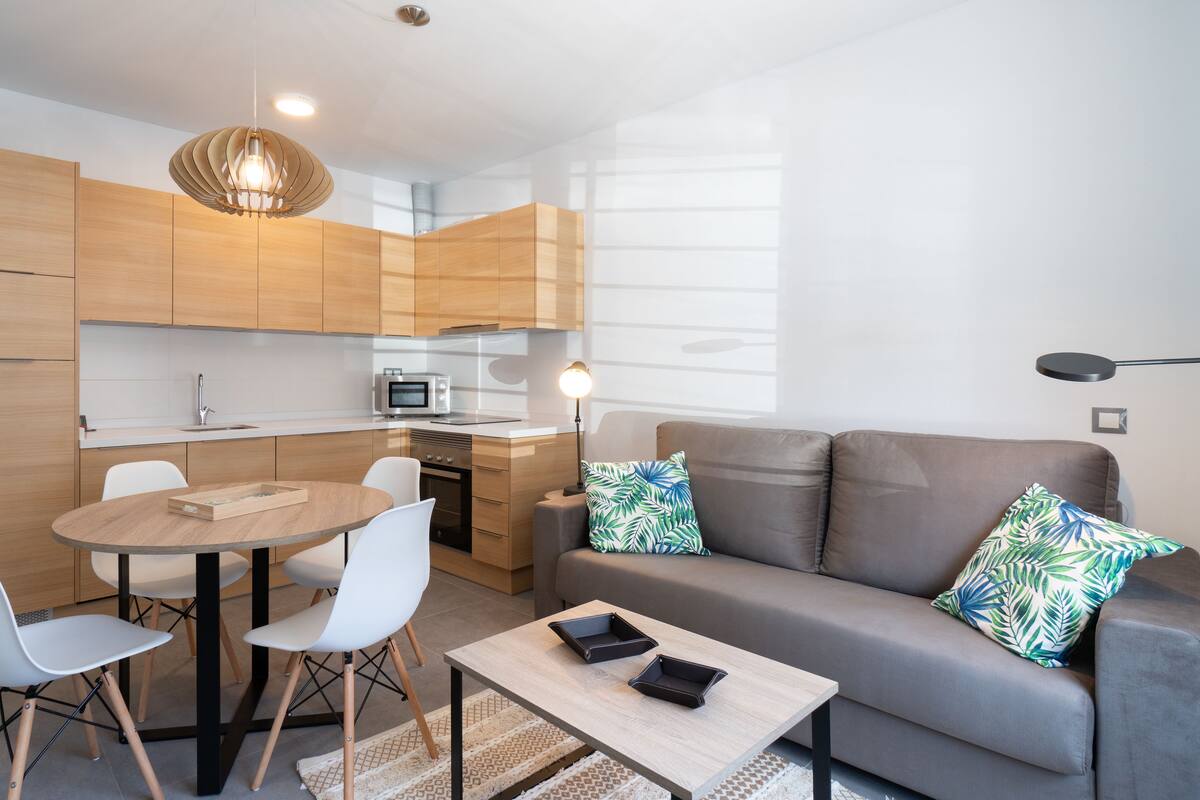 A modern kitchen with wooden cabinets, a dining table with chairs, a sofa with decorative pillows, and a coffee table.