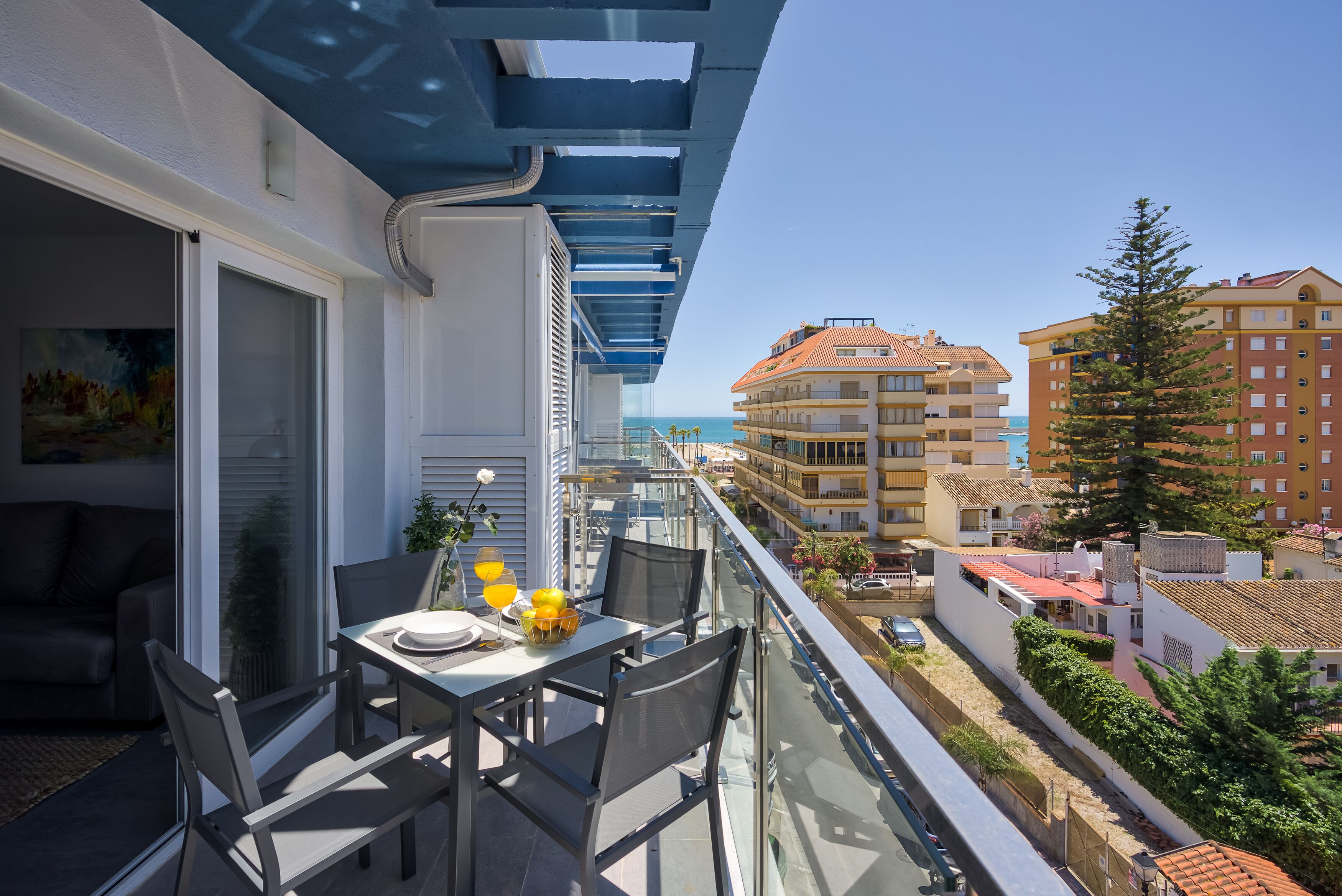 A balcony with a table and chairs, overlooking a coastal area with buildings and a clear sky.