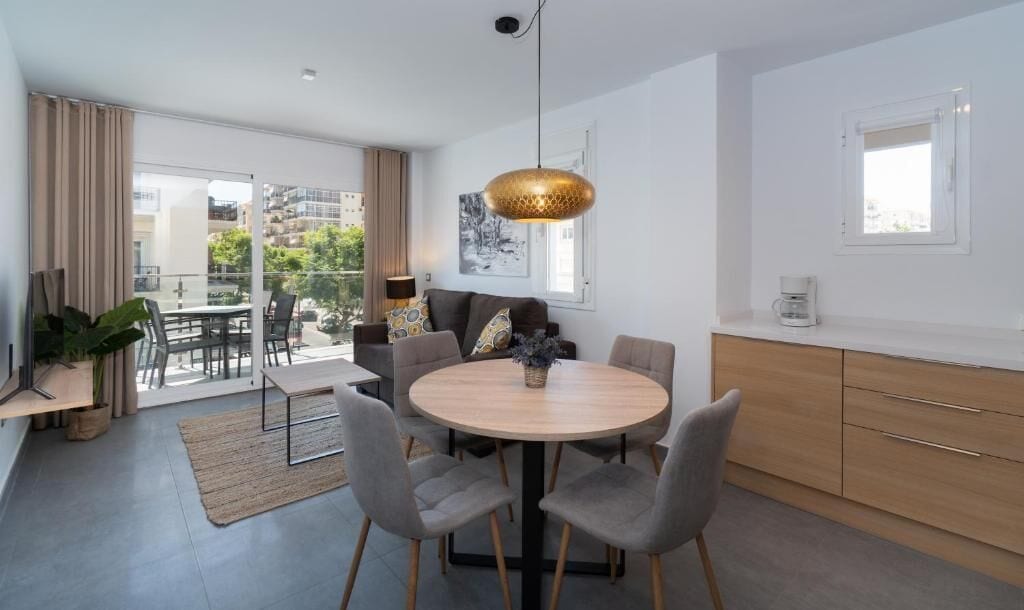 Un salon moderne avec une table ronde en bois, des chaises grises, un canapé agrémenté de coussins à motifs, une suspension et une vue sur l’extérieur grâce à de grandes fenêtres.