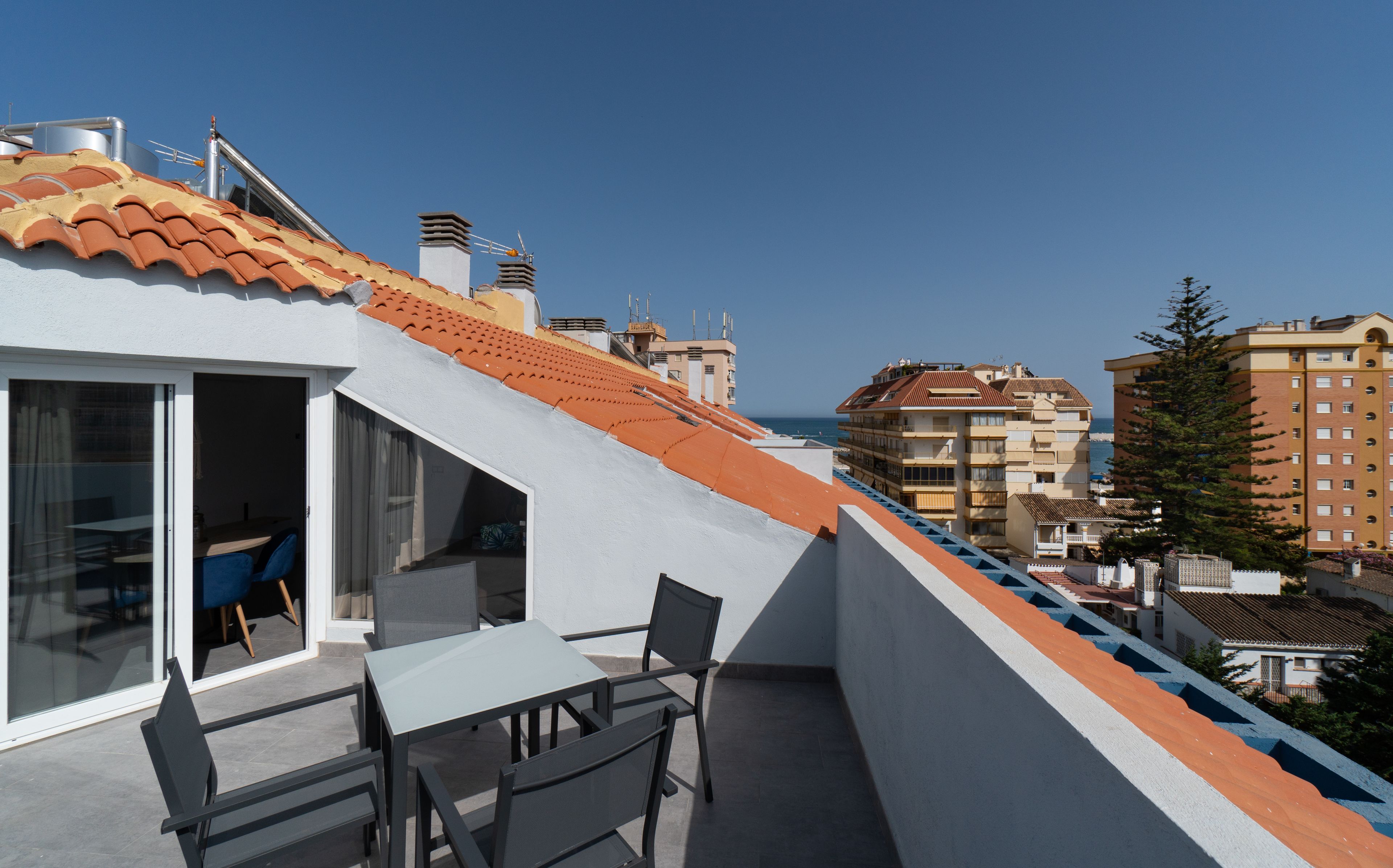 A balcony with a table and chairs, overlooking a coastal area with buildings and a clear sky.