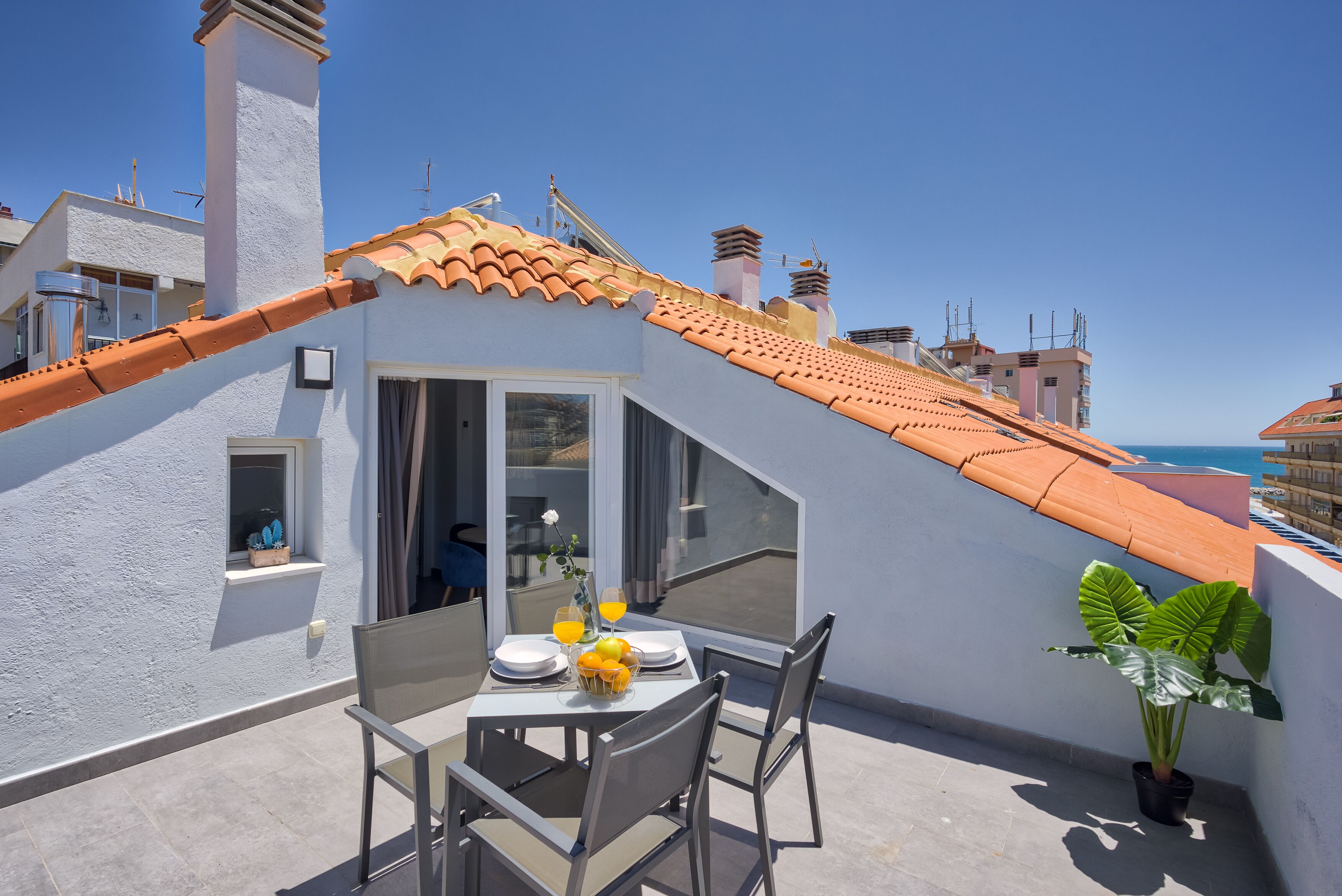 A rooftop terrace with a table set for two, a potted plant, and a view of the sea.
