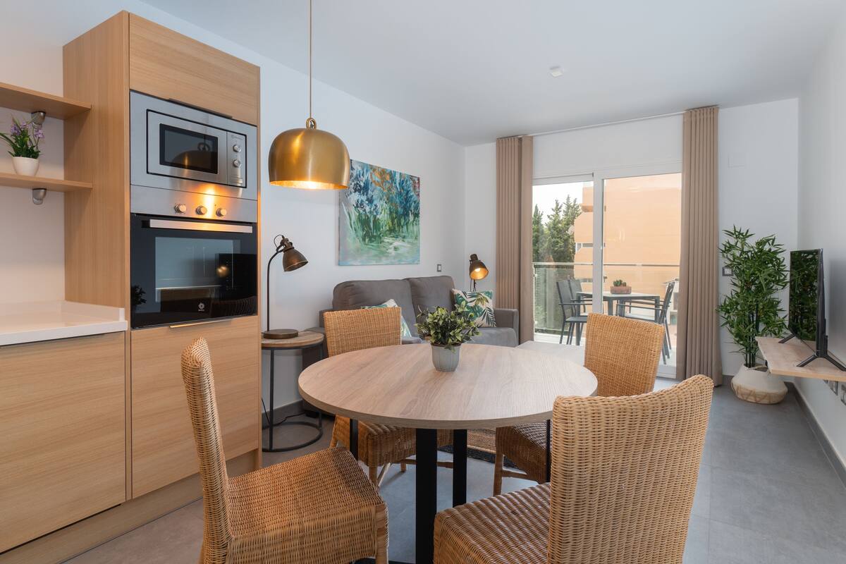 A modern kitchen with built-in appliances, a dining area with wicker chairs, and a balcony with a view.