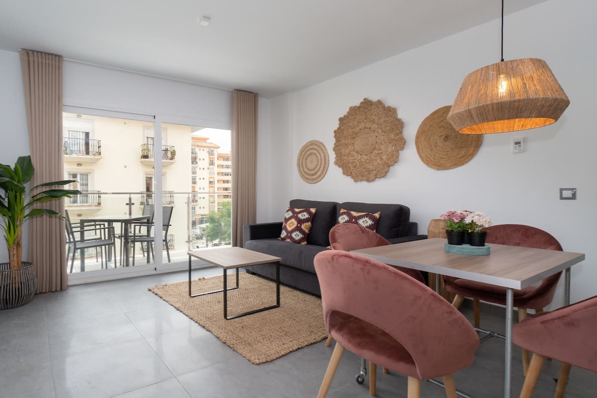 A modern living room with a sofa, dining table, chairs, a TV, and a balcony with a view of buildings.